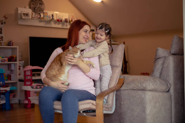 The pregnant woman is sitting on a chair with the cat in her lap. The little girl is standing on the chair, and she wants to hold the cat, too.